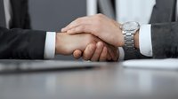 Two businessmen in formal suits shaking hands across office desk, symbolizing successful partnership, agreement, and professional collaboration in corporate environment.