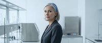 Mature woman with gray hair and fair complexion wearing dark business blazer standing in modern laboratory or office environment with scientific equipment and glass partitions visible in background.
