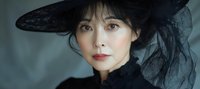 Young Asian woman in elegant black wide-brimmed hat with tulle veil detail, wearing dark clothing against blurred background, dramatic fashion portrait in moody lighting.