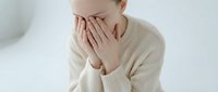 Young woman covering face with hands showing stress, anxiety or depression in beige sweater against white background for mental health concepts.