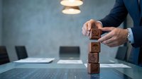 Professional businessman in suit stacking wooden blocks with icons in modern office environment representing business strategy and growth planning.