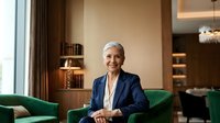 Mature woman with gray hair and light skin tone, wearing professional navy blazer and white blouse, seated on green velvet chair in modern luxury office or hotel lobby with warm lighting and contemporary furnishings.