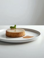 Round chocolate mousse dessert with mint leaf garnish and caramel sauce served on white ceramic plate against neutral background, minimalist food presentation.