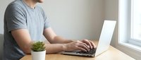 Man in gray t-shirt typing on laptop at wooden desk with small potted plant, working from home office space near window.