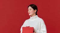 Young Asian woman in white turtleneck sweater holding red gift box against vibrant red background, perfect for holiday or celebration themes.