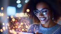 Young African American woman with curly hair and glasses smiling while holding magical glowing particles in her hands against blurred city lights background.