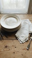Stacked cream ceramic plates with embossed rim detail, white linen napkin with lace trim, and vintage silverware arranged on rustic wooden table near window for dining and tableware styling.