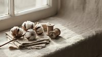 Cotton bolls with wooden spools of thread and knitting needles on linen fabric by window creating rustic crafting scene for handmade textile projects.