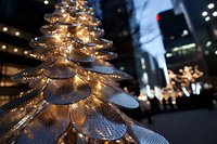 Modern metallic Christmas tree sculpture with glowing lights against urban cityscape background, creating festive downtown atmosphere at night.