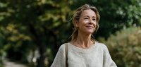Mature Caucasian woman with gray hair smiling outdoors in autumn park wearing beige sweater. Natural lifestyle portrait for wellness and aging concepts.