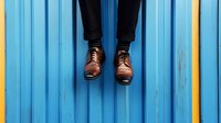 Person in dark jeans and brown leather shoes sitting on blue wooden bench or deck surface for casual lifestyle and fashion concepts.