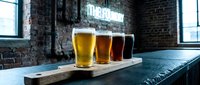 Four glasses of craft beer in varying shades from light to dark arranged on a wooden serving board in an industrial brewery or pub setting with exposed brick walls.