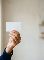 Adult hand holding blank white card against neutral wall background, wearing dark blue sweater sleeve. Mockup for business cards, invitations, or promotional materials.