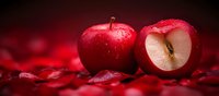 Fresh red apples with water drops on dark background with rose petals, one apple cut in half showing juicy flesh and seed, romantic food styling composition.