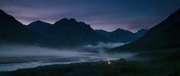 Tent glowing with warm light in misty mountain valley at dusk, surrounded by dramatic peaks and fog-covered landscape for camping and adventure themes.