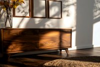 Wooden mid-century modern sideboard in sunlit room with tree shadow patterns on wall, empty frames and autumn leaves in vase creating cozy atmosphere.
