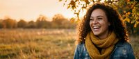 Happy young African American woman with curly hair wearing mustard scarf and denim jacket laughing outdoors during golden hour sunset in autumn field.