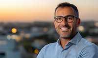 Middle-aged Mediterranean businessman wearing glasses and blue shirt smiles confidently against blurred urban sunset background with bokeh lights, expressing success and leadership.