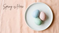Pastel Easter eggs on a white plate over peach linen with Spring is Here handwritten text, flat lay holiday greeting card.