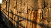 Weathered concrete wall with rust stains and dramatic shadow patterns from metal railing creating geometric lines on textured urban surface.