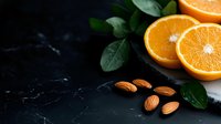 Fresh halved oranges with raw almonds and green leaves on dark marble background, moody food photography composition for healthy eating concept.