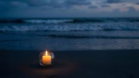 Glowing candle in glass jar on sandy beach at twilight with ocean waves and dramatic cloudy sky creating peaceful meditation atmosphere for relaxation concepts.