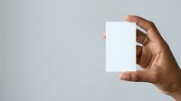 Male hand with medium brown skin tone holding blank white card or paper against neutral gray background for branding and marketing mockups.