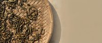 Dried tea leaves in woven basket on neutral background, overhead view for beverage and wellness design projects.