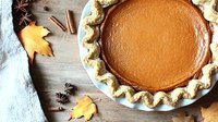 Fresh baked pumpkin pie with decorative fluted crust on rustic wooden table with autumn maple leaves, cinnamon sticks and star anise spices.