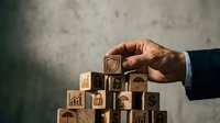 Businessman hand placing wooden block with shield icon on stack of cubes with business symbols for protection and security concept.