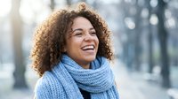 Young mixed-race woman with curly hair laughing joyfully wearing blue knit scarf in urban winter setting. Authentic happiness and positive emotions concept.