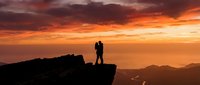 Silhouette of couple standing on rocky cliff edge at sunset with golden orange sky and distant mountains, romantic landscape scenery for travel and lifestyle use.