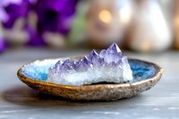 Purple amethyst crystal cluster in ceramic bowl on wooden surface with blurred flowers background for meditation and healing practices.