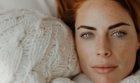 Close up portrait of young Caucasian woman with natural freckles, blue eyes and red hair wearing white knitted sweater, looking at camera with gentle smile.