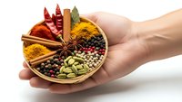 Hands holding wooden bowl with colorful spices including turmeric, chili, cardamom, star anise and peppercorns arranged in compartments for cooking.