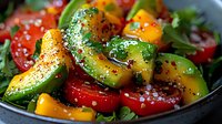 Fresh summer salad with juicy tomatoes, ripe avocado chunks, and mango pieces garnished with black pepper and herbs in dark bowl. Healthy vegetarian meal.