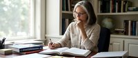 Mature businesswoman with glasses writing notes at home office desk near window with bookshelves, professional remote work environment for corporate planning.