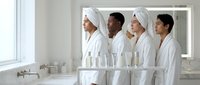 Group of diverse young adults wearing white bathrobes and towel wraps in modern bathroom, looking at mirror reflection during spa or wellness routine.