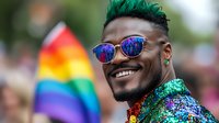 Young African American man with green hair and sequined jacket smiling at Pride parade celebration, wearing reflective sunglasses with rainbow flag in background. LGBTQ+ community event.