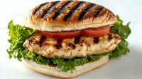 Grilled chicken sandwich with fresh lettuce, tomato slices on toasted bun showing grill marks and seasoned meat. Close-up food photography on light background.