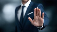 Businessman in navy suit making stop gesture with palm forward, representing boundary setting, rejection, or limit establishment in professional environment.