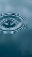 Water droplet creating concentric ripples on calm blue surface with soft bokeh background for wellness and meditation concepts.