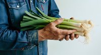 Farmer in denim shirt holding fresh green leeks with roots, isolated on white background for agricultural and food industry use.