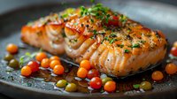Grilled salmon fillet with herbs, cherry tomatoes and olives on dark plate, close up view of cooked fish with colorful garnish in savory sauce.