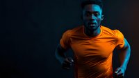 Young African American male athlete in orange t-shirt running against dark background with dramatic blue lighting effect creating dynamic sports atmosphere.