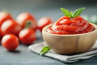 Fresh tomato sauce in wooden bowl garnished with basil leaf, whole red tomatoes in background on gray textile napkin, selective focus macro shot.