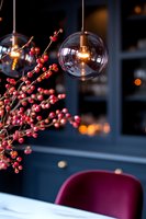 Modern glass pendant lamp with glowing filament bulb against dark blue wall, red berry branches and burgundy chair creating cozy autumn atmosphere.