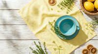 Turquoise ceramic cup on yellow linen napkin with fresh thyme, dried lemon slices, and chamomile on rustic white wooden table.