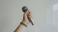 Hand holding wireless microphone with chain bracelet against neutral background for broadcasting and audio recording applications.