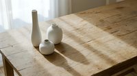 Minimalist ceramic vases in white tones arranged on wooden table with natural sunlight casting shadows, suitable for home decor and interior design projects.
